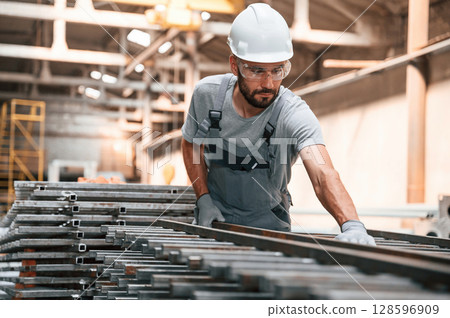Taking care of metal products. Young factory worker in grey uniform Taking care of metal products. Young factory worker in grey uniform 128596909