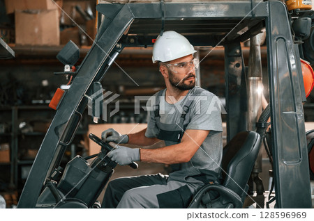 Driving the forklift. Young factory worker in grey uniform Driving the forklift. Young factory worker in grey uniform 128596969