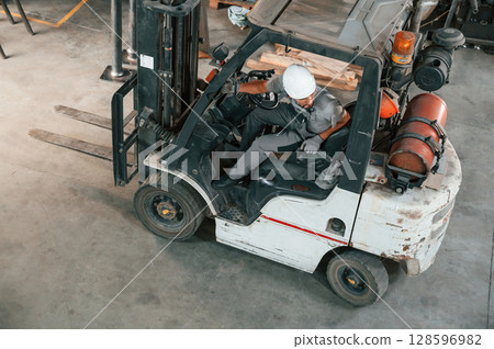 View from the top, in the forklift. Young factory worker in grey uniform View from the top, in the forklift. Young factory worker in grey uniform 128596982