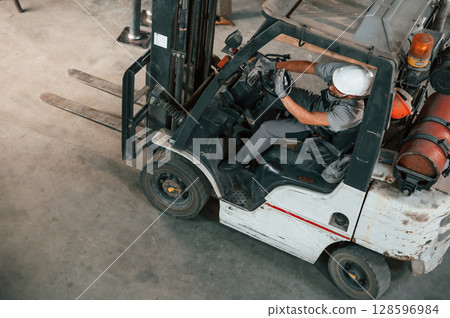 View from the top, in the forklift. Young factory worker in grey uniform View from the top, in the forklift. Young factory worker in grey uniform 128596984