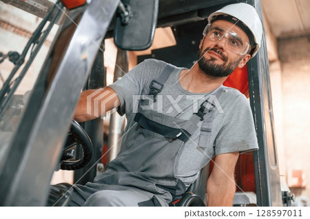 In the forklift. Young factory worker in grey uniform In the forklift. Young factory worker in grey uniform 128597011