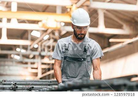 In glasses and white hard hat. Young factory worker in grey uniform 128597018