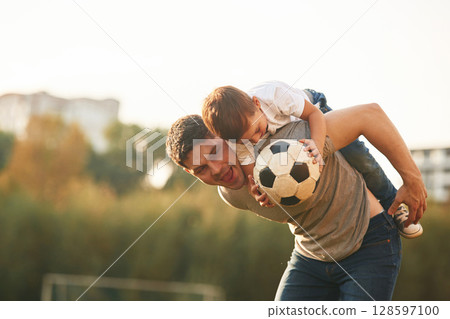 Holding professional soccer ball. Father and little son are playing and having fun outdoors 128597100