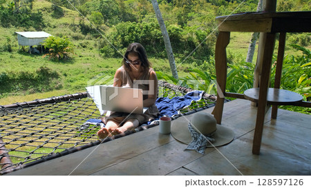 A young woman enjoys her freelance work from a hammock, nestled on a tropical terrace, surrounded by lush rice fields and vibrant jungle A young woman enjoys her freelance work from a hammock, nestled on a tropical terrace, surrounded by lush rice fields and vibrant jungle 128597126