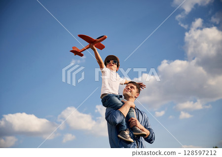 Playing with toy plane, sitting on the shoulders. Father and little son are having fun outdoors 128597212