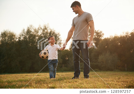 Walking and holding soccer ball. Father and little son are playing and having fun outdoors Walking and holding soccer ball. Father and little son are playing and having fun outdoors 128597257