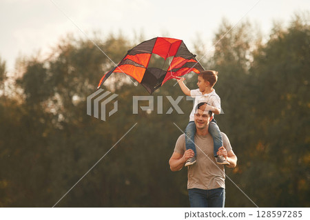 Red and black colored kite in hands. Father and little son are playing and having fun outdoors Red and black colored kite in hands. Father and little son are playing and having fun outdoors 128597285