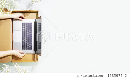 Buying and delivering new laptop. Romantic overhead shot of womans hands holding laptop in cardboard box on blue background with white spring flowers. Flat lay, copy space Buying and delivering new laptop. Romantic overhead shot of womans hands holding laptop in cardboard box on blue background with white spring flowers. Flat lay, copy space 128597342