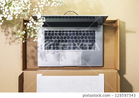 Buying and delivering new technics. Romantic sustainable overhead shot of laptop in cardboard box on cardboard background with white spring flowers and shadows. Flat lay 128597343