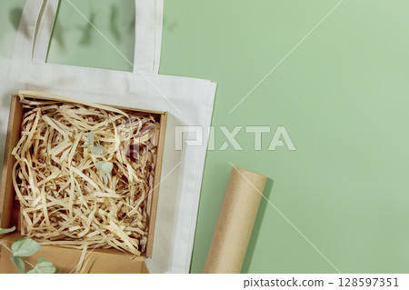 Eco friendly packaging concept. Cardboard box with shredded paper on textile shopping bag on green background with green leaves. Eco packaging goods from online stores and delivery. Copy space 128597351