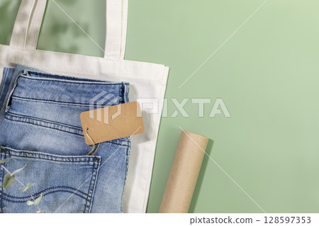 Sustainable still life with jeans with empty craft label, textile bag and packing paper. Eco friendly clothing shop. Casual clothes store, shopping, sale and denim shop concept. Flat lay, copy space Sustainable still life with jeans with empty craft label, textile bag and packing paper. Eco friendly clothing shop. Casual clothes store, shopping, sale and denim shop concept. Flat lay, copy space 128597353