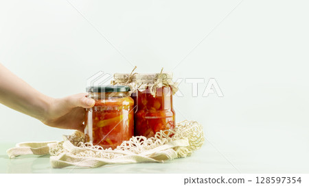 Probiotic foods. Sustainable still life with woman hand hold pickled or fermented vegetables. Lecho and ajvar in glass jars in mesh bag on blue background. Home food preserving or canning. Copy space Probiotic foods. Sustainable still life with woman hand hold pickled or fermented vegetables. Lecho and ajvar in glass jars in mesh bag on blue background. Home food preserving or canning. Copy space 128597354