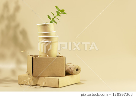 Sustainable still life. Eco friendly packaging concept. Cardboard boxes, disposable cups with green shoot on beige background. Concept of ecology, conservation, plastic free and recycling. Copy space 128597363