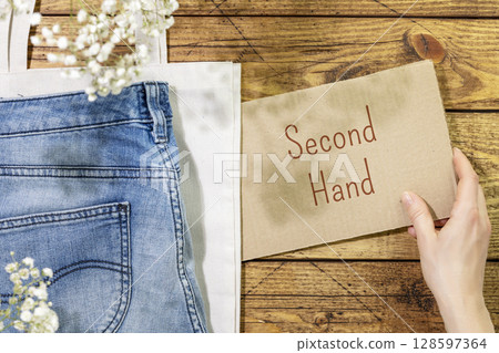 Sustainable still life with jeans on textile bag and female hand holding inscription second hand. Second hand shop. Circular fashion, eco friendly sustainable shopping, thrifty shop concept. Flat lay 128597364