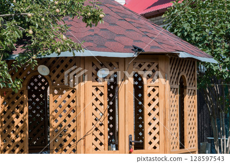 Rustic Wooden Gazebo with Red Shingle Roof Amidst Lush Greenery, Outdoor Living, Garden Scene, Countryside Home, Summer Sunlight, Natural Ambiance. Rustic Wooden Gazebo with Red Shingle Roof Amidst Lush Greenery, Outdoor Living, Garden Scene, Countryside Home, Summer Sunlight, Natural Ambiance. 128597543