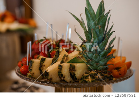Vibrant Fruit Platter Display with Pineapple, Strawberries, Oranges, and Skewers on Elegant Glass Stand, Bright Tropical Color Palette. Vibrant Fruit Platter Display with Pineapple, Strawberries, Oranges, and Skewers on Elegant Glass Stand, Bright Tropical Color Palette. 128597551