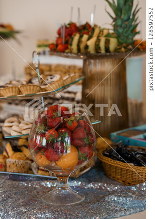 Elegant Dessert Table with Fresh Fruit Display, Pastries, Strawberries, Pineapple, Gold Accents, Silver Tablecloth, Party Buffet Setup. Elegant Dessert Table with Fresh Fruit Display, Pastries, Strawberries, Pineapple, Gold Accents, Silver Tablecloth, Party Buffet Setup. 128597552