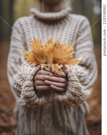 A woman in a cozy knit sweater gently holds a bunch of yellow leaves in a forest, celebrating fall fashion and seasonal warmth 128598062
