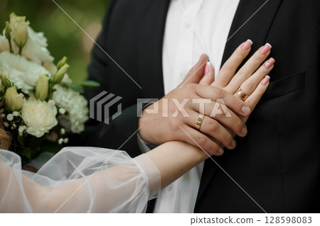 Wedding Rings on Hands of Groom and Bride, Close-Up Shot, White Bouquet, Classic Gold Bands, Marriage, Love, Romance, Formal Attire, Soft Focus 128598083