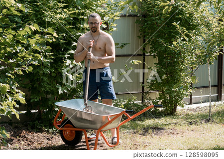 Shirtless Man Gardening with Wheelbarrow Outdoors, Green Bushes, Sunny Day, Natural Setting, Backyard, Orange Handle, Agricultural Work, Summer. 128598095