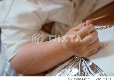 Baby's Hand Clasping Gold Necklace Chain on White Textile with Intricate Beadwork Detail, Close-Up, Soft Lighting Baby's Hand Clasping Gold Necklace Chain on White Textile with Intricate Beadwork Detail, Close-Up, Soft Lighting 128598141