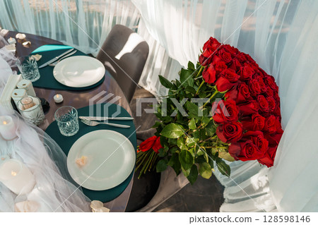 Romantic Dinner Setting with Red Roses: Luxurious Tablecloth, Elegant Plates, Cozy Candles, and Intimate Ambience in a Bright, Airy Space. 128598146