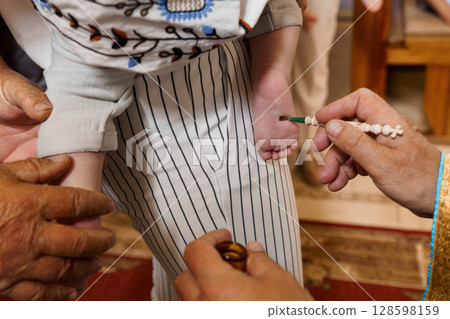 Traditional Henna Foot Ritual: Close-up of Baby's Sole with Intricate Floral Design, Hands Applying Paste, Warm Indoor Setting, Brown and Beige Tones 128598159