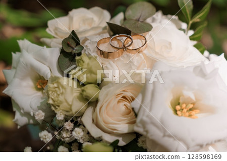 Wedding Rings on White Bouquet. Romantic Marriage Symbolism, Rose Flowers, Gold Bands, Blurred Forest Background, Delicate Floral Arrangement, Love. Wedding Rings on White Bouquet. Romantic Marriage Symbolism, Rose Flowers, Gold Bands, Blurred Forest Background, Delicate Floral Arrangement, Love. 128598169
