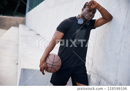 Resting by leaning on the white wall. Young black man is with basketball ball outdoors Resting by leaning on the white wall. Young black man is with basketball ball outdoors 128599106