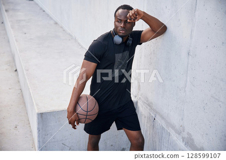 Resting by leaning on the white wall. Young black man is with basketball ball outdoors 128599107