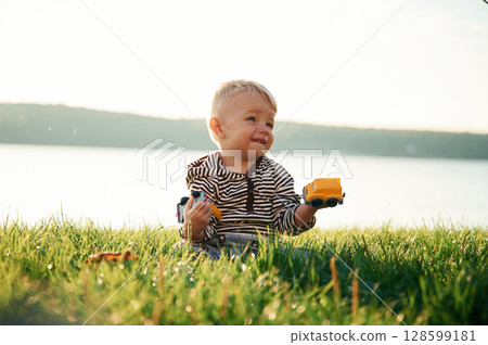Against lake behind. Cute little boy is having fun outdoors on the ground with green grass 128599181