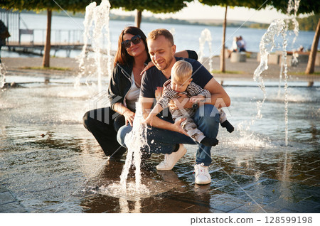 Family of father, mother and little boy are outdoors, enjoying the water from fountain Family of father, mother and little boy are outdoors, enjoying the water from fountain 128599198