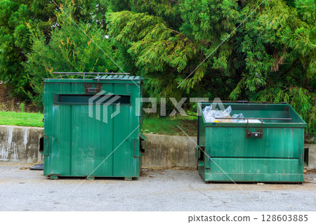 Two green dumpsters sit side by side outside, surrounded by trees grass on sunny day. 128603885