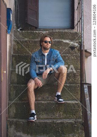 A young man in a denim jacket and white T-shirt sits on outdoor stairs, smiling 128604106