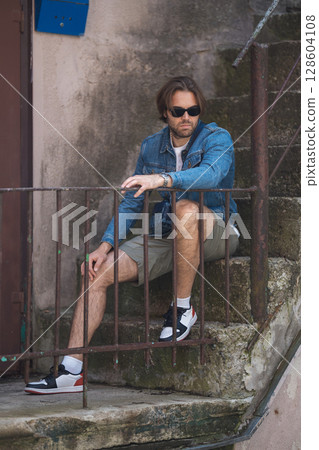 A young man in a denim jacket and white T-shirt sits on outdoor stairs, smiling A young man in a denim jacket and white T-shirt sits on outdoor stairs, smiling 128604108