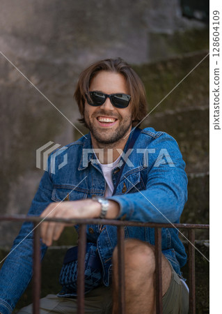 A young man in a denim jacket and white T-shirt sits on outdoor stairs, smiling 128604109