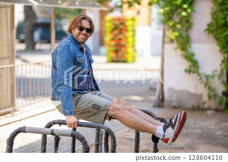 A stylish young man in a denim jacket, white T-shirt, shorts, and sneakers poses 128604110