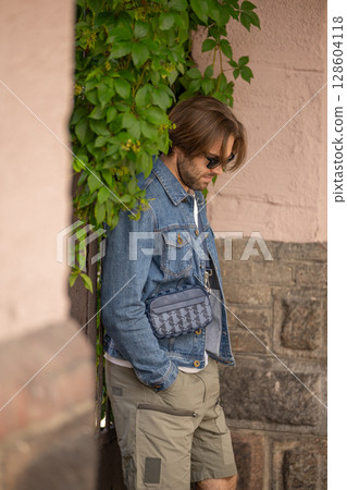A stylish young man in a denim jacket and sunglasses poses confidently against a rustic wall A stylish young man in a denim jacket and sunglasses poses confidently against a rustic wall 128604118