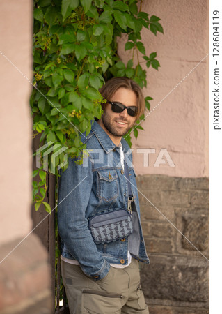 A stylish young man in a denim jacket and sunglasses poses confidently against a rustic wall A stylish young man in a denim jacket and sunglasses poses confidently against a rustic wall 128604119