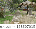 Tropical stone path leading through a lush garden with frangipani trees and fallen blossoms. Features a large thatched umbrella, outdoor lights, and rustic seating areas, evoking a serene resort. 128607015