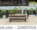 Rustic wooden bench on gravel ground, surrounded by potted plants. Background features a building with a balcony, wooden railing, and chairs, hinting at a cozy outdoor cafe or resort setting. 128607016