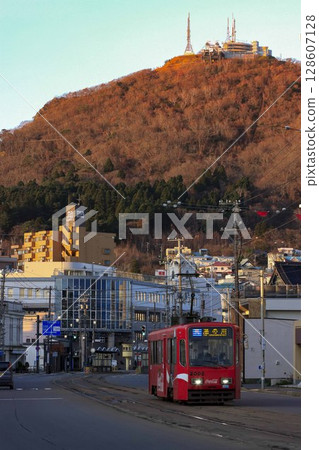 [Vertical composition, logo and driver blurred] Hakodate City Tram running with the autumn leaves of Mount Hakodate in the background, Hakodate City 128607128