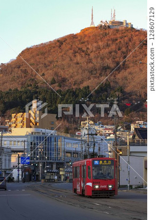 [Vertical composition, logo and driver blurred] Hakodate City Tram running with the autumn leaves of Mount Hakodate in the background, Hakodate City 128607129