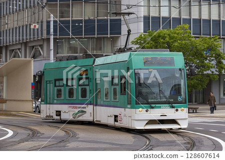 [Partially edited, blurred driver logo] Hakodate City Tram passing through the curve in front of Hakodate Station 128607614