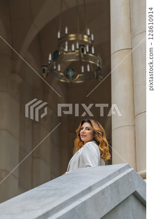 Woman Chandelier Castle - A woman in a white dress stands on a stone staircase in a grand castle hallway with a chandelier above. 128610914