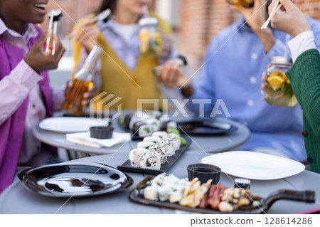 Friends enjoying sushi outdoors, sharing food and drinks at a cafe or restaurant. 128614286