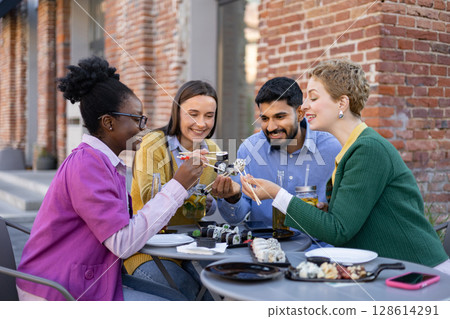 A diverse group of friends enjoys sushi outdoors, sharing food and laughter around a table. A diverse group of friends enjoys sushi outdoors, sharing food and laughter around a table. 128614291