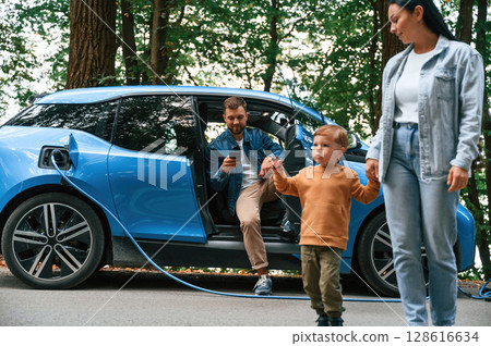 Walking forward. Mother, father and little son are waiting for electric car to charge outdoors 128616634