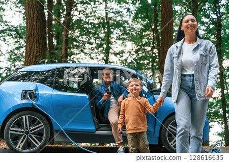 Walking forward. Mother, father and little son are waiting for electric car to charge outdoors 128616635