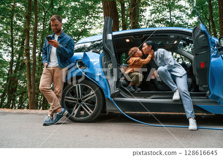 Conception of wellness. Mother, father and little son are waiting for electric car to charge outdoors Conception of wellness. Mother, father and little son are waiting for electric car to charge outdoors 128616645